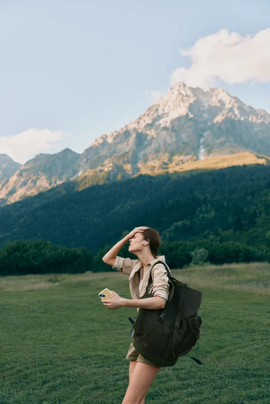 Woman hiking outdoors with backpack, enjoying nature and mountain scenery, sunlit field, casual apparel, sense of adventure, travel, relaxed lifestyle, fresh air.