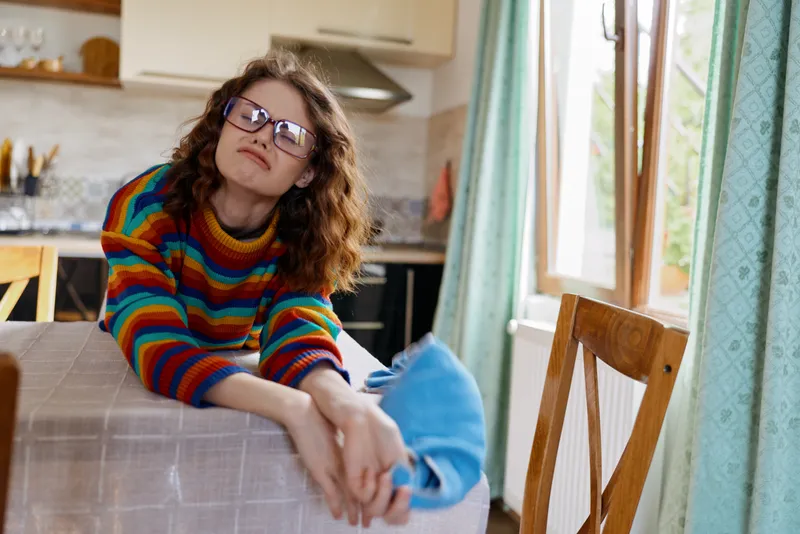 A young woman with curly brunette hair and sunglasses leans over a wooden kitchen table, wearing a colorful striped sweater, smiling softly in a bright, cozy home setting.