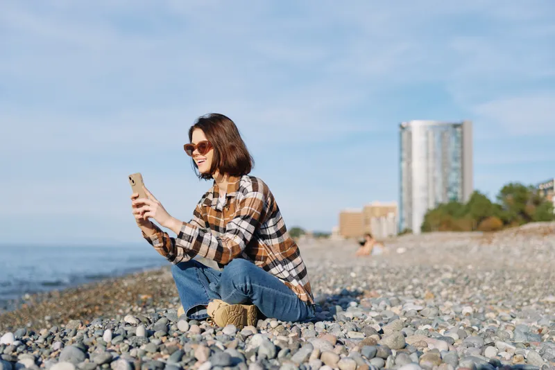 Woman on a pebbled beach selfies with phone under sunny skies, wearing casual plaid shirt and jeans, sunglasses, outdoor mood by the coast and sea