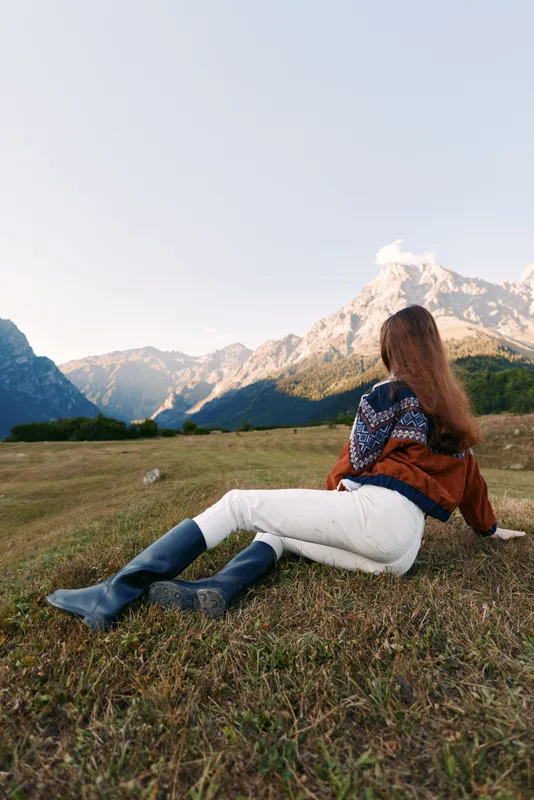 Woman in a colorful sweater and boots sitting on a meadow facing towering mountains, enjoying nature and panoramic landscape with relaxation, solitude and autumn light.