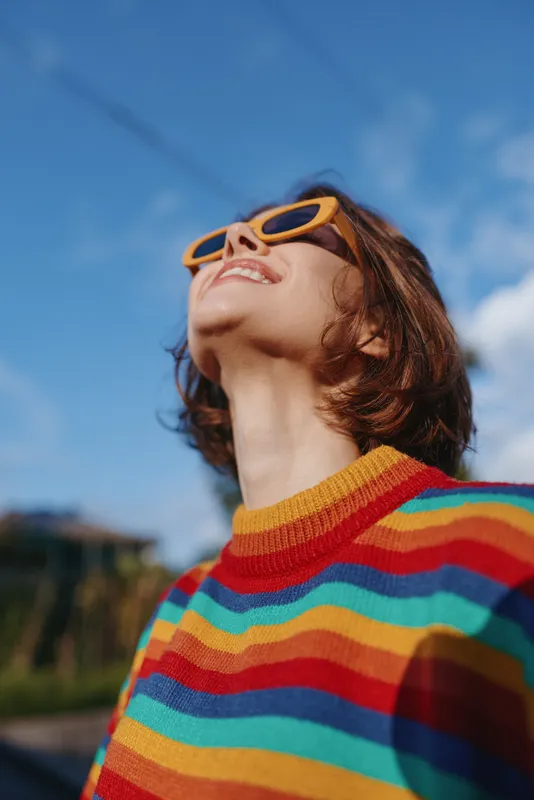 Woman smiling in a colorful striped sweater and orange sunglasses, looking up with short brown hair against a clear blue sky outdoors; casual outfit, joyful travel lifestyle portrait.