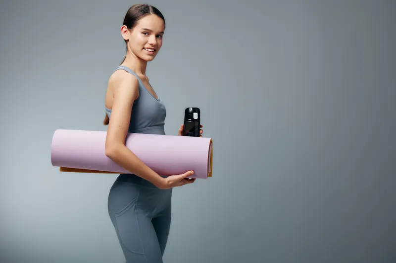 Young woman in athletic wear carries a yoga mat and water bottle, ready for a workout. A clean studio backdrop highlights fitness, sport and wellness in motion.