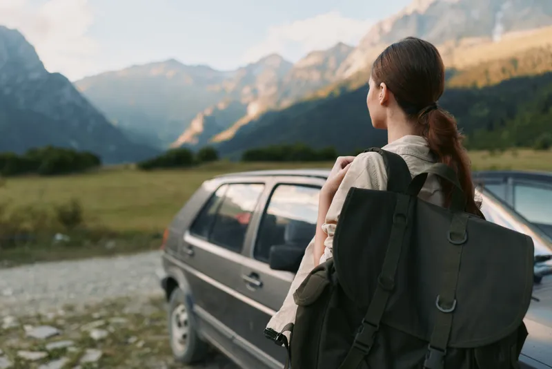 Woman traveler with backpack standing near car looking at mountain landscape on sunny day, outdoor adventure, nature exploration, casual lifestyle.