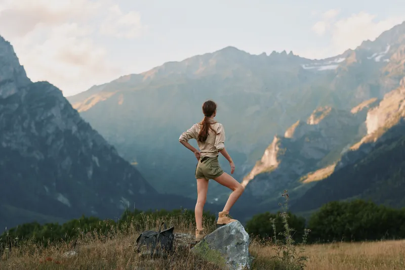 Young woman standing on a rock overlooking dramatic mountains, exploring the outdoors in hiking shorts. A lifestyle scene that conveys nature, adventure, and wellness.