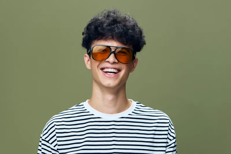 Smiling young man wearing striped shirt and oversized orange sunglasses against a plain olive background, showing happiness and casual style.