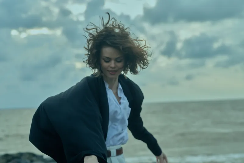 Woman on a windy beach in a white shirt and dark jacket moving forward, sea and clouds behind, conveying freedom, motion, outdoor fashion and dramatic atmosphere today.