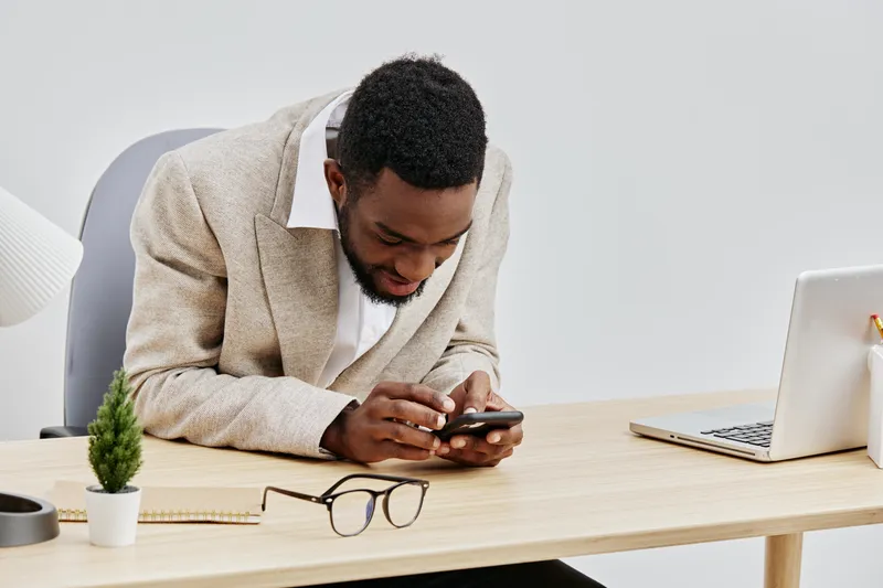 Modern African American man sitting at desk using smartphone in bright office environment with minimal decor, laptop, glasses, and plant, working or browsing on mobile device, professional lifestyle c