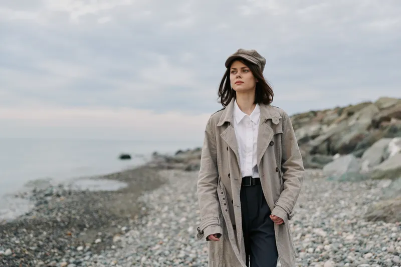 Young woman in stylish coat and hat walks on rocky beach under cloudy sky. Outdoor fashion portrait with natural landscape and serene mood. Concept of autumn style and casual elegance.