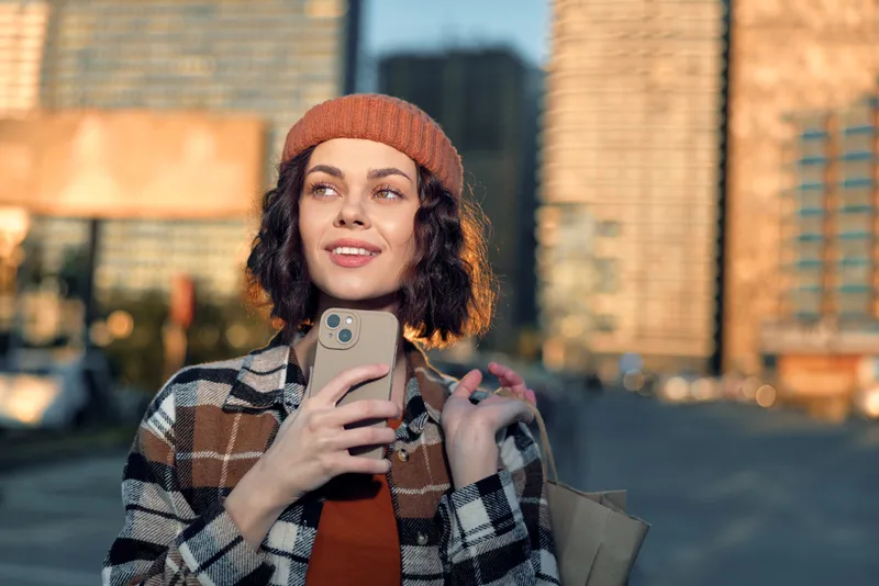 Woman in city holding smartphone and shopping bag, smile and beanie in autumn light, candid lifestyle portrait with golden hour glow, authenticity and mindful living vibe.
