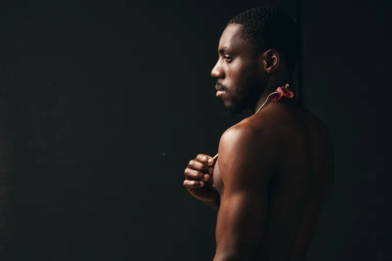 Creative visual designer style portrait of African American man with dark skin and flower on shoulder, side view with bare upper body on black background in studio lighting.