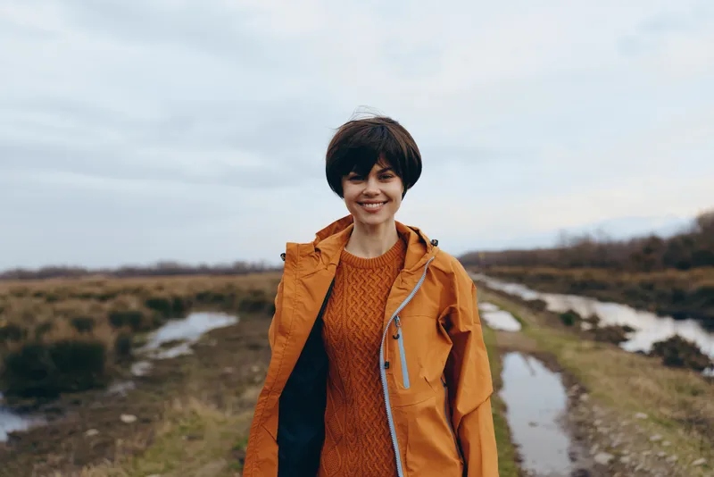 Smiling woman outdoors in countryside, orange jacket and knit sweater, standing near a dirt path, winter landscape with snow patches, lifestyle concept, natural daylight, relaxed mood