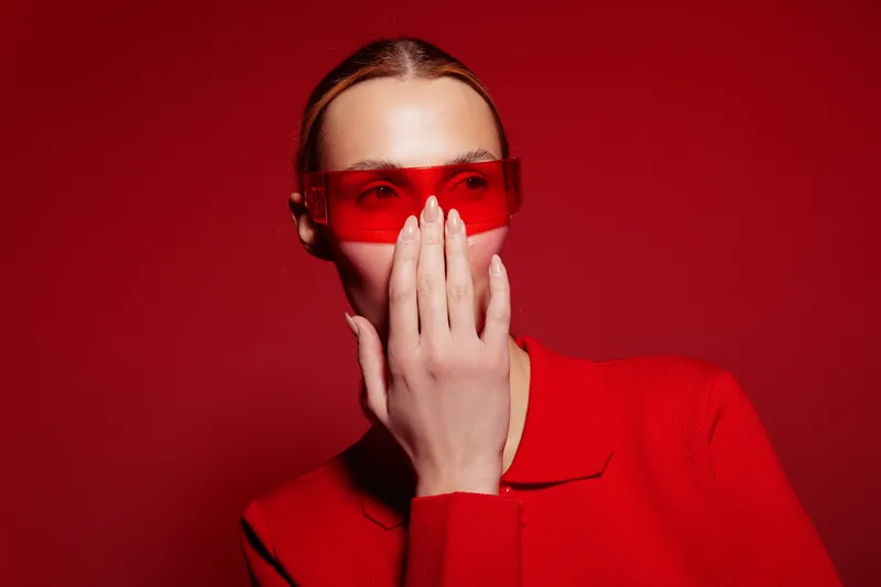 Young woman in red sweater and red sunglasses covers mouth with hand against red background. Concept of fashion, mystery, and modern style with monochrome color scheme.