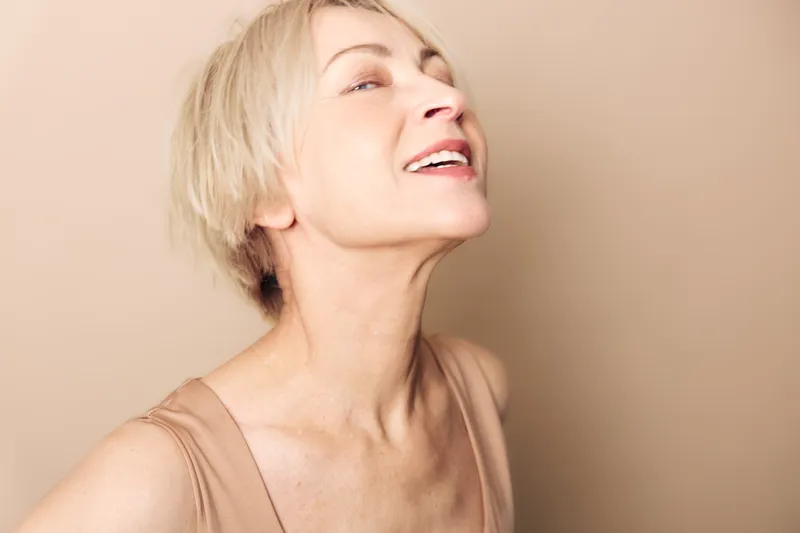 Smiling mature woman with short blonde hair looks upward in beige tank top against light background. Concept of confidence, happiness, natural beauty, and aging gracefully.