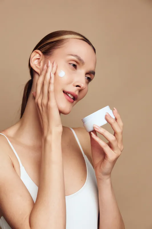 Young woman with blonde hair applies white face cream on her cheek while holding a jar in her other hand against a neutral background, expressing calm and self-care.
