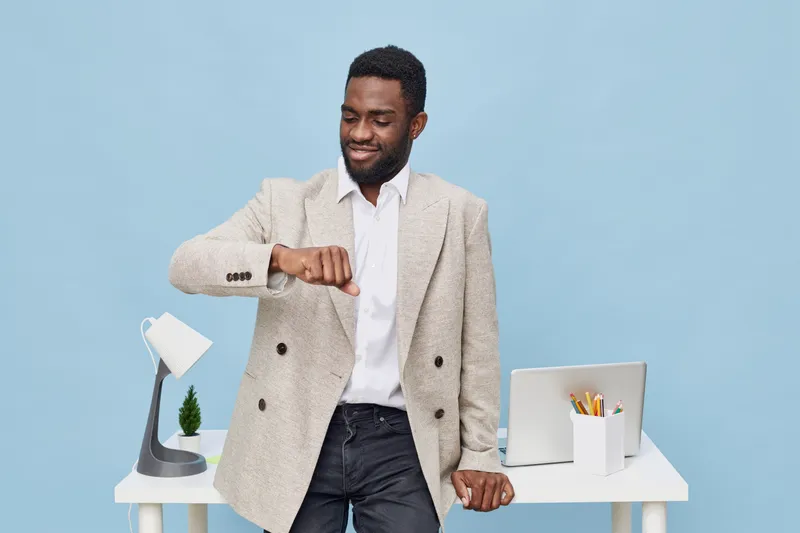 Confident young African American man in smart casual outfit checking watch while standing at modern workspace with laptop, desk accessories, and blue background. Business and time management concept.