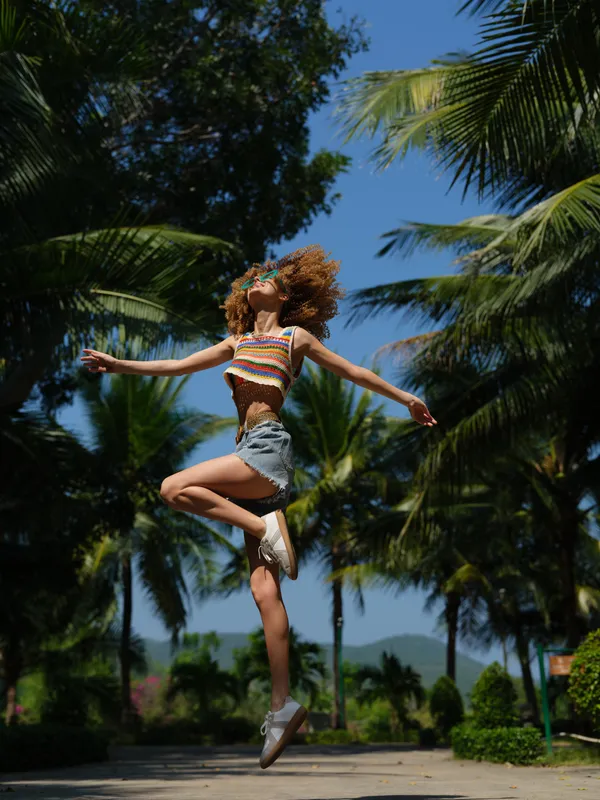 energetic young woman jumping outdoors in casual summer clothes, enjoying sunny day with palm trees and clear blue sky in background, active lifestyle concept