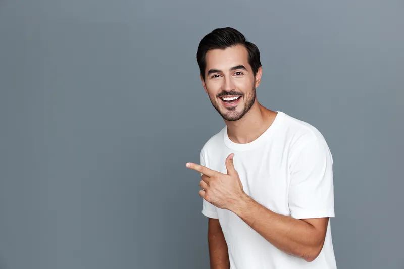 Cheerful smiling young man in casual white t-shirt pointing finger while looking at the camera on gray studio background. Positive lifestyle concept.