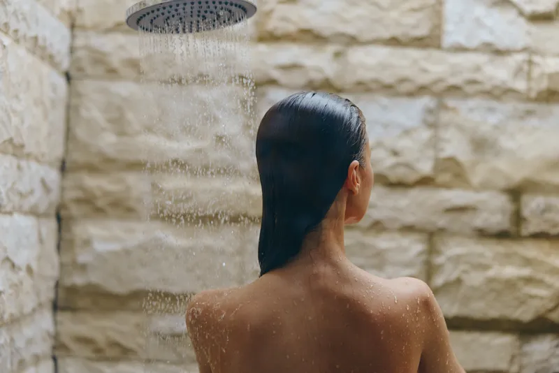 Woman enjoying outdoor shower with stone wall background, wet hair, and water droplets cascading down her back in a serene natural setting.