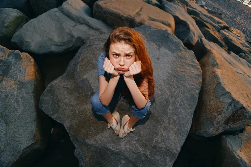 young woman sitting on large rocks outdoors with serious expression wearing casual clothes and sandals under sunlight in natural rocky environment