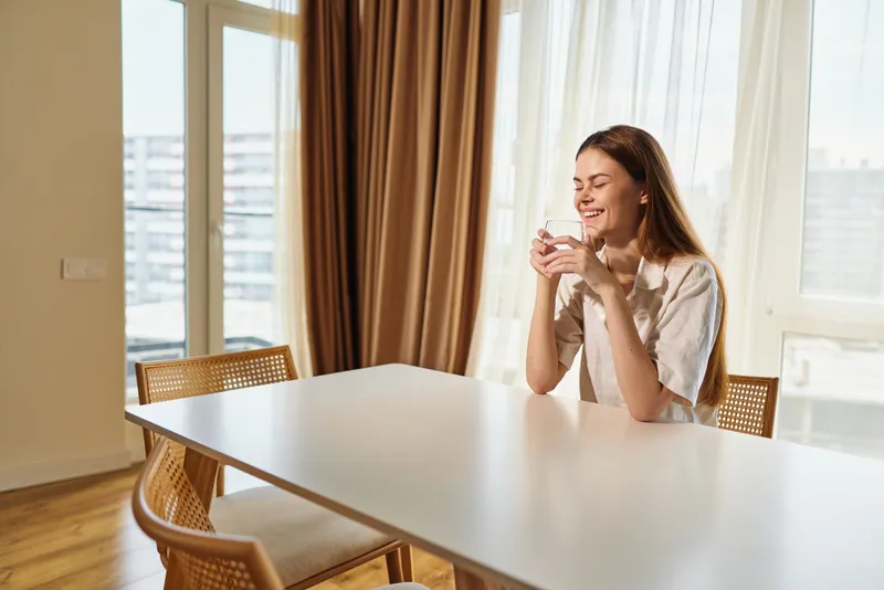 Woman smiling with cup at table in bright home morning sunlight by window, enjoying coffee or tea. Relaxed female portrait in cozy dining interior, casual clothing and peaceful moment.