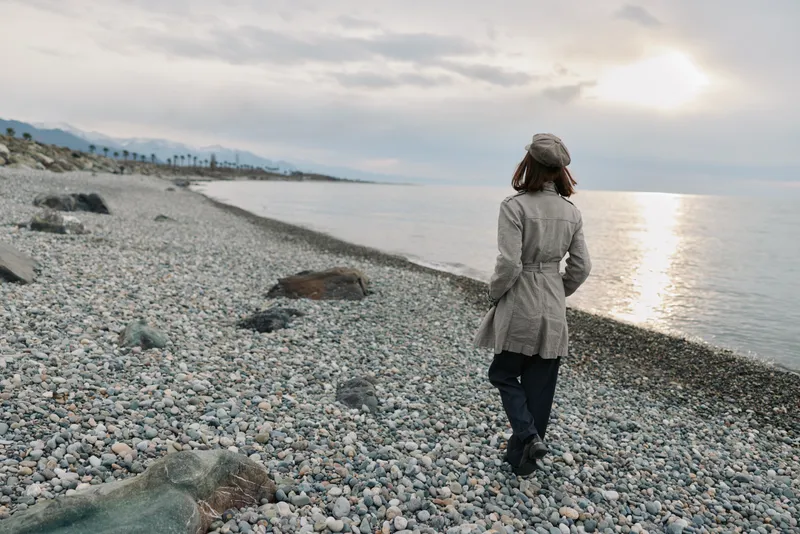 Woman in coat and hat walks alone on a pebble beach near calm sea during cloudy sunset. Concept of solitude, nature, and peaceful outdoor moment by the water.