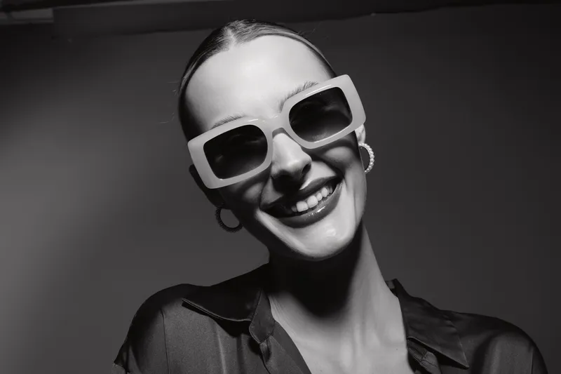 Smiling white woman with light skin wearing large square sunglasses and hoop earrings enjoys studio portrait in black and white. Concept of fashion, style, and confidence.