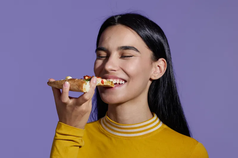 woman sport fitness enjoying slice of pizza with closed eyes in casual yellow outfit against purple background, healthy balance concept