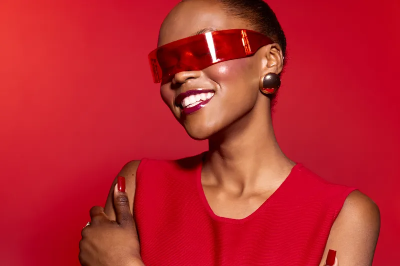 Smiling black woman with dark skin wearing red dress and futuristic red glasses poses confidently on red background. Concept for fashion, beauty, and modern style with vibrant color.