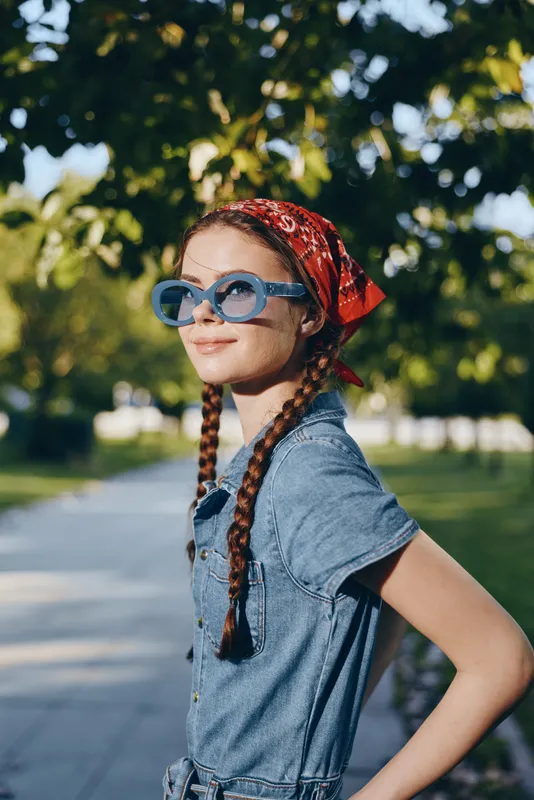 Lifestyle woman wearing modern boho-western denim outfit with blue sunglasses and red bandana, styled in film color, outdoors on sunny day in relaxed street style setting.