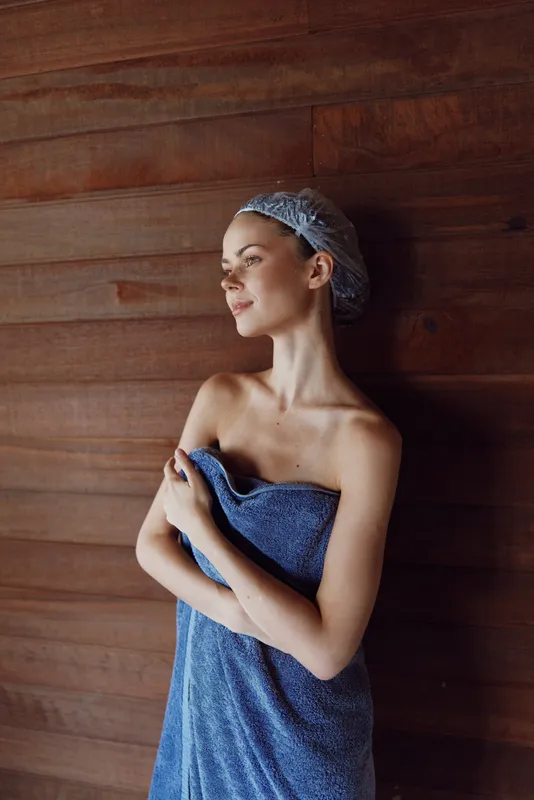 Young woman wrapped in a blue towel with a hair cap, standing against a wooden wall, showing calm and relaxed expression in natural light indoors.