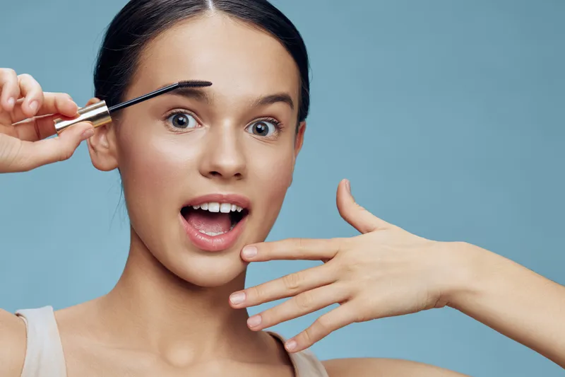 Young woman applying mascara with excitement, showcasing a beauty routine against a light blue background, emphasizing freshness and self care