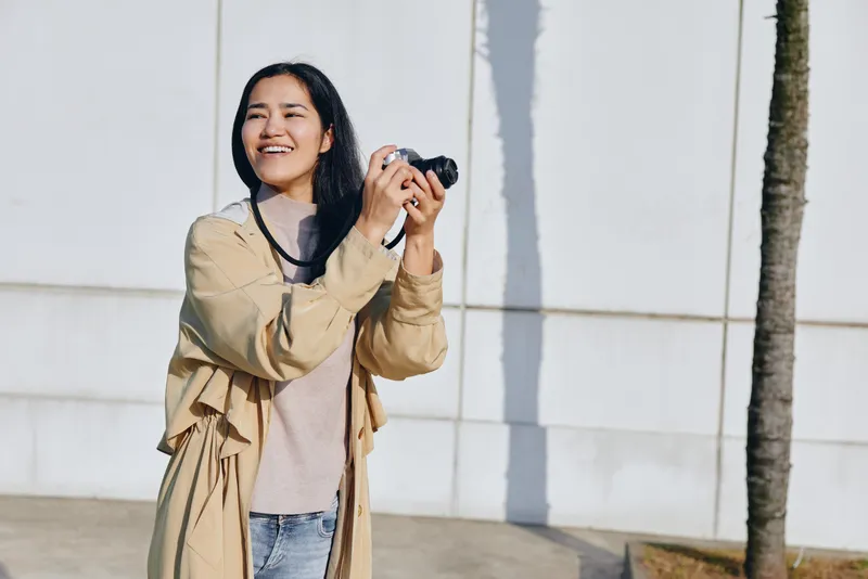 Woman holding a camera outdoors, smiling beside a white wall and palm tree, wearing a beige trench and light sweater, showcasing casual fashion and street photography vibe