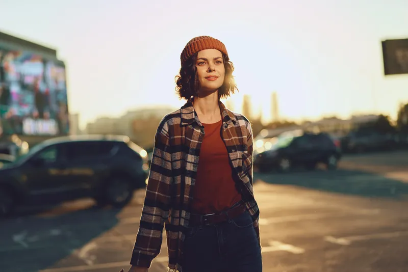 Woman portrait in urban parking lot at golden hour, candid lifestyle shot of smiling beanie model in plaid jacket, authenticity and emotional storytelling with warm golden hour glow.