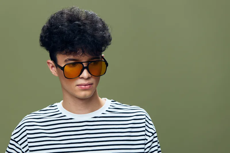 Young man wearing striped shirt and amber sunglasses against green background, curly black hair, casual style, thoughtful expression, studio portrait.