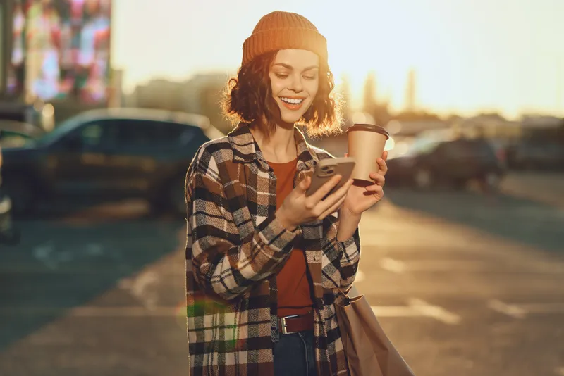 Woman with coffee and phone smiling on urban street at golden hour, candid lifestyle portrait in beanie and plaid coat, authenticity and emotional storytelling for mindful living.
