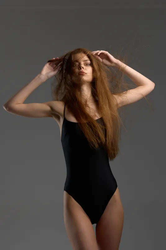 Young woman with long hair in black swimsuit posing confidently against gray background.