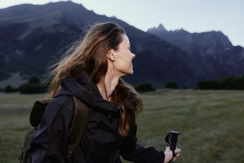 A lone hiker woman stands in a rugged landscape beneath distant mountains, gazing to the side with a calm smile, wearing a dark jacket and backpack, embodying outdoor adventure.