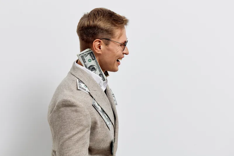 Professional portrait of smiling man with glasses, wearing beige blazer and white shirt, standing against plain light background. Casual business style, positive expression, confident demeanor, studio