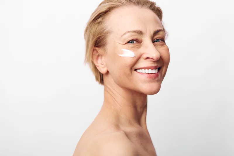 Smiling mature woman with clear skin applies white facial cream on cheek in bright studio. Concept for skincare, anti aging, natural beauty, wellness, and self care with positive mood.