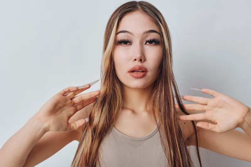Young woman with long straight hair and neutral facial expression posing on white background. Beauty model, casual style, natural makeup and light sleeveless top in studio setting.