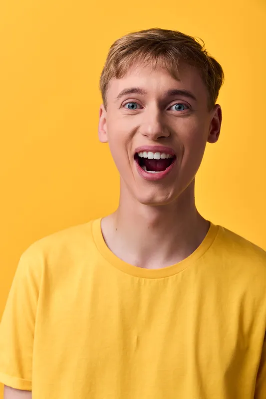 Smiling, youthful man in a bright yellow shirt against a vibrant background conveys happiness, energy and casual style. Closeup shot highlights cheerful expression, warm lighting and modern fashion