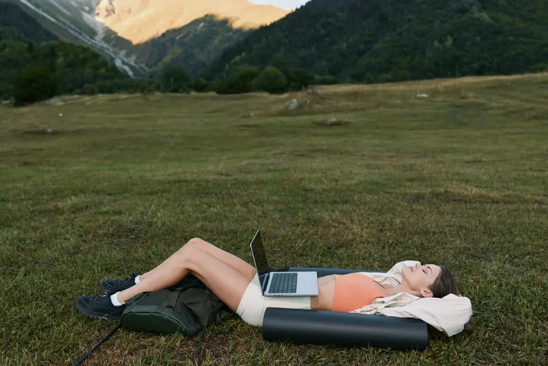 A person lies on a portable air couch in an open field, laptop resting on their stomach, enjoying a peaceful outdoor work break and a moment of calm in nature.