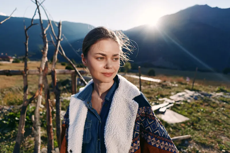 Woman portrait in mountain outdoors at countryside with sunlight, smiling in warm patterned jacket. Travel and nature scene with serene expression and rustic landscape background.