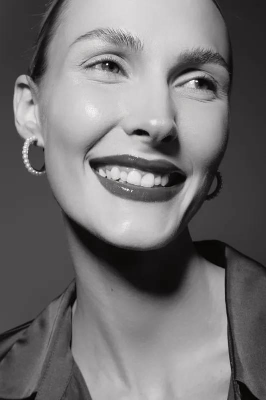 Closeup portrait of a smiling Caucasian woman with fair skin and hoop earrings. The black and white photo captures natural beauty and joyful expression in studio lighting.