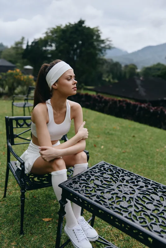 A young woman in sporty attire, sitting outdoors on a chair, exuding a contemplative emotion while surrounded by greenery and mountains, perfect for fitness themes