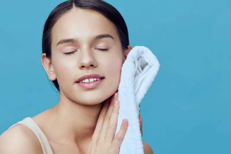 Young female model gently caring for her skin with a towel, showcasing healthy skincare, happiness, and beauty against a bright blue background