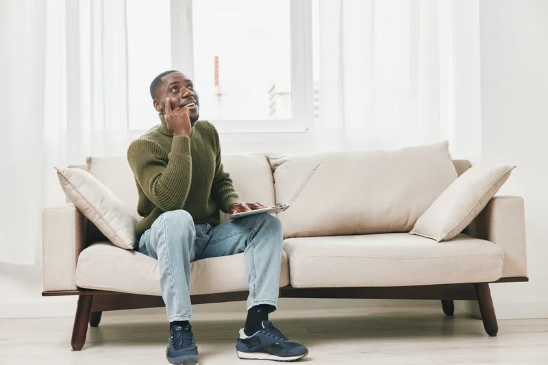 Thoughtful Black man working on a laptop while sitting on a comfortable couch in a stylish room, contemplating ideas and productivity