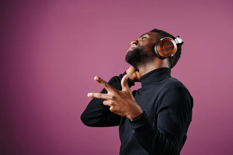 Young Black man enjoying music with headphones, wearing a stylish black turtleneck, expressing joy against a vibrant pink background