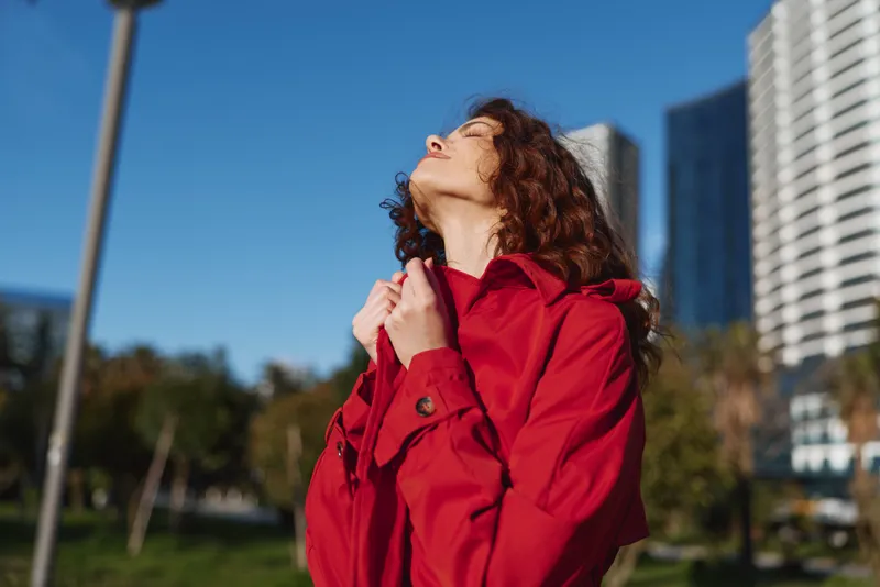 Woman in a red coat outdoors, basking in sunlight, blue sky above urban buildings, relaxed pose, natural colors, candid city portrait against a backdrop