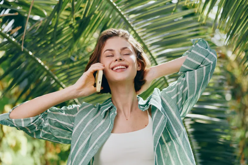 A cheerful woman stands outdoors among palm leaves, smiling while talking on a mobile phone, wearing a striped shirt.